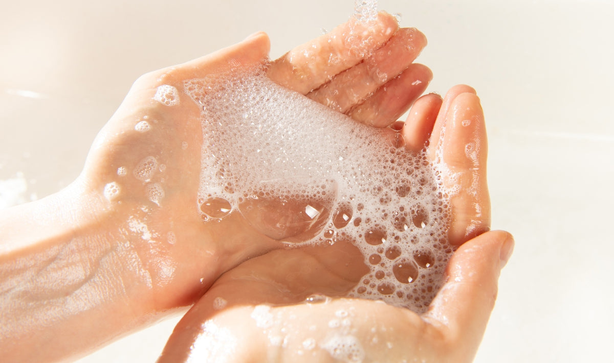 Deux mains tenant des bulles de savon blanches et mousseuses sous une lumière vive, évoquant le lavage des mains ou la propreté.