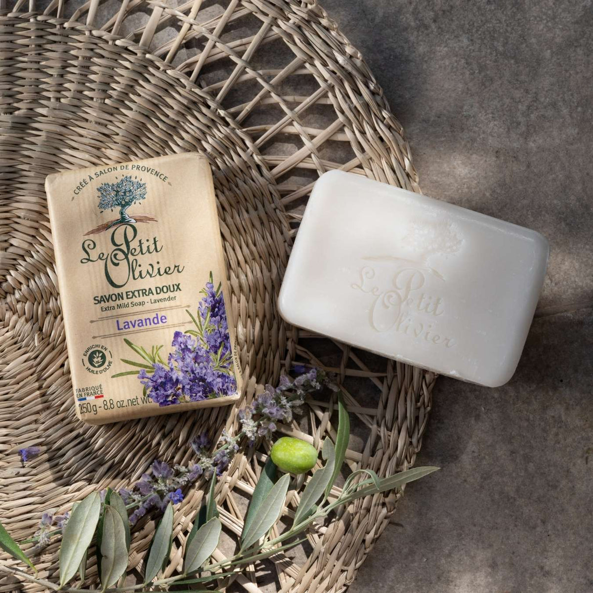 Un savon à la lavande « Le Petit Olivier » et son emballage reposent sur une natte tressée, accompagnés de brins de lavande et d'un rameau d'olivier, baignés d'une douce lumière naturelle.
