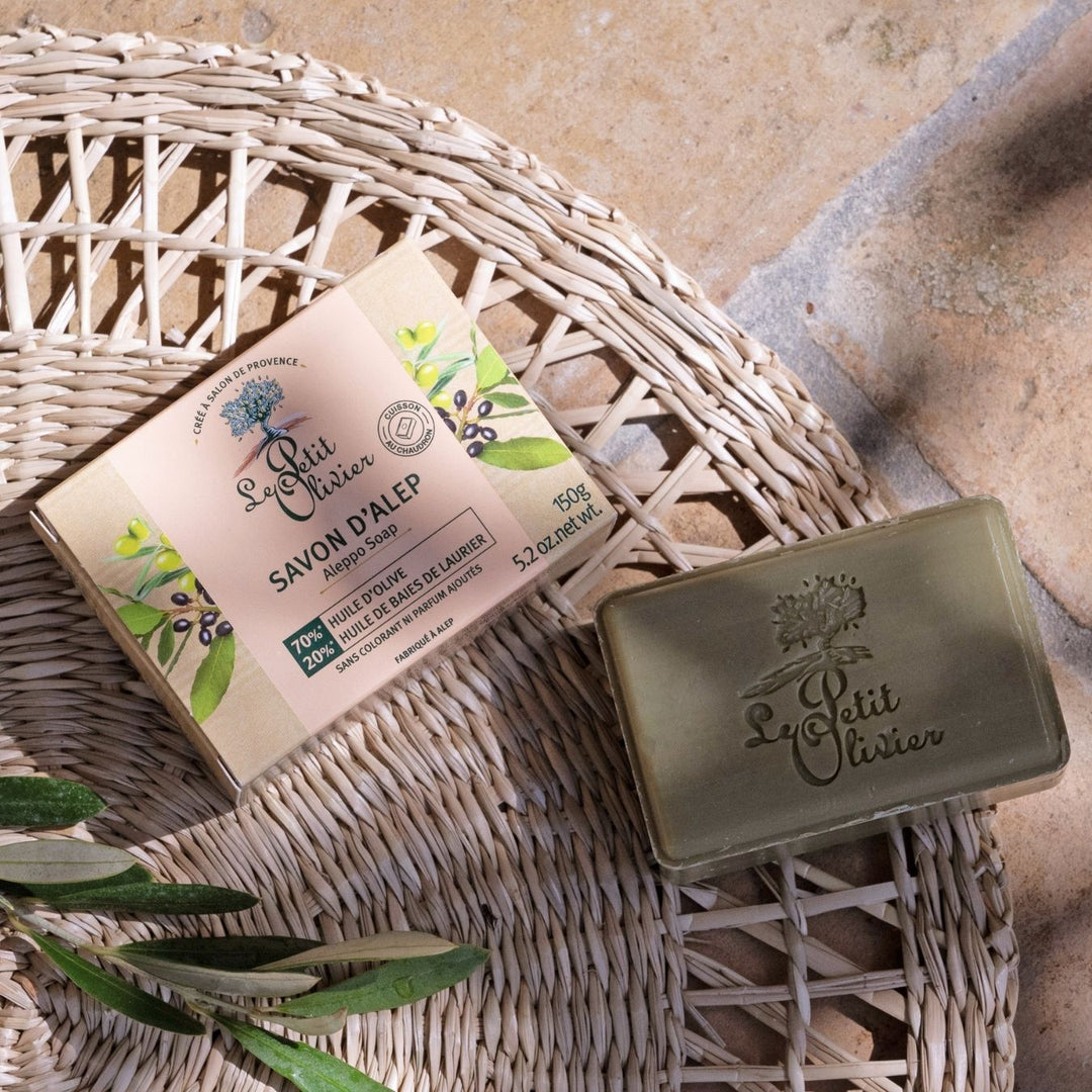 Un savon « Le Petit Olivier Savon d'Alep », à base d'huile d'olive, repose sur un support tressé, baigné par la lumière du soleil, à côté de son coffret et d'un rameau d'olivier — idéal pour les peaux sensibles.