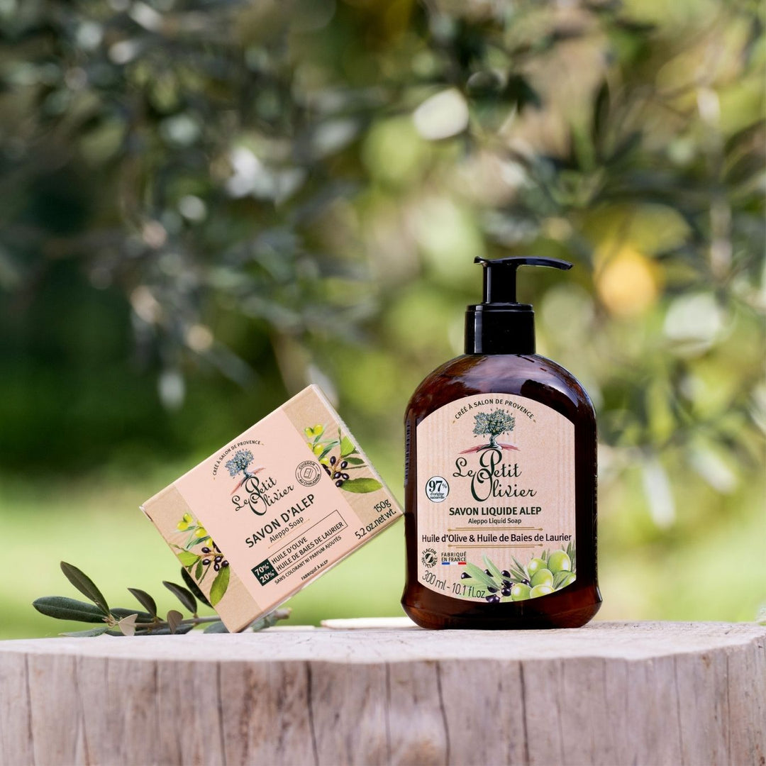 Le savon d’Alep « Le Petit Olivier », enrichi en huile d’olive, repose à l’air libre sur un support en bois, au milieu de la verdure et des branches d’olivier — idéal pour les peaux sensibles.