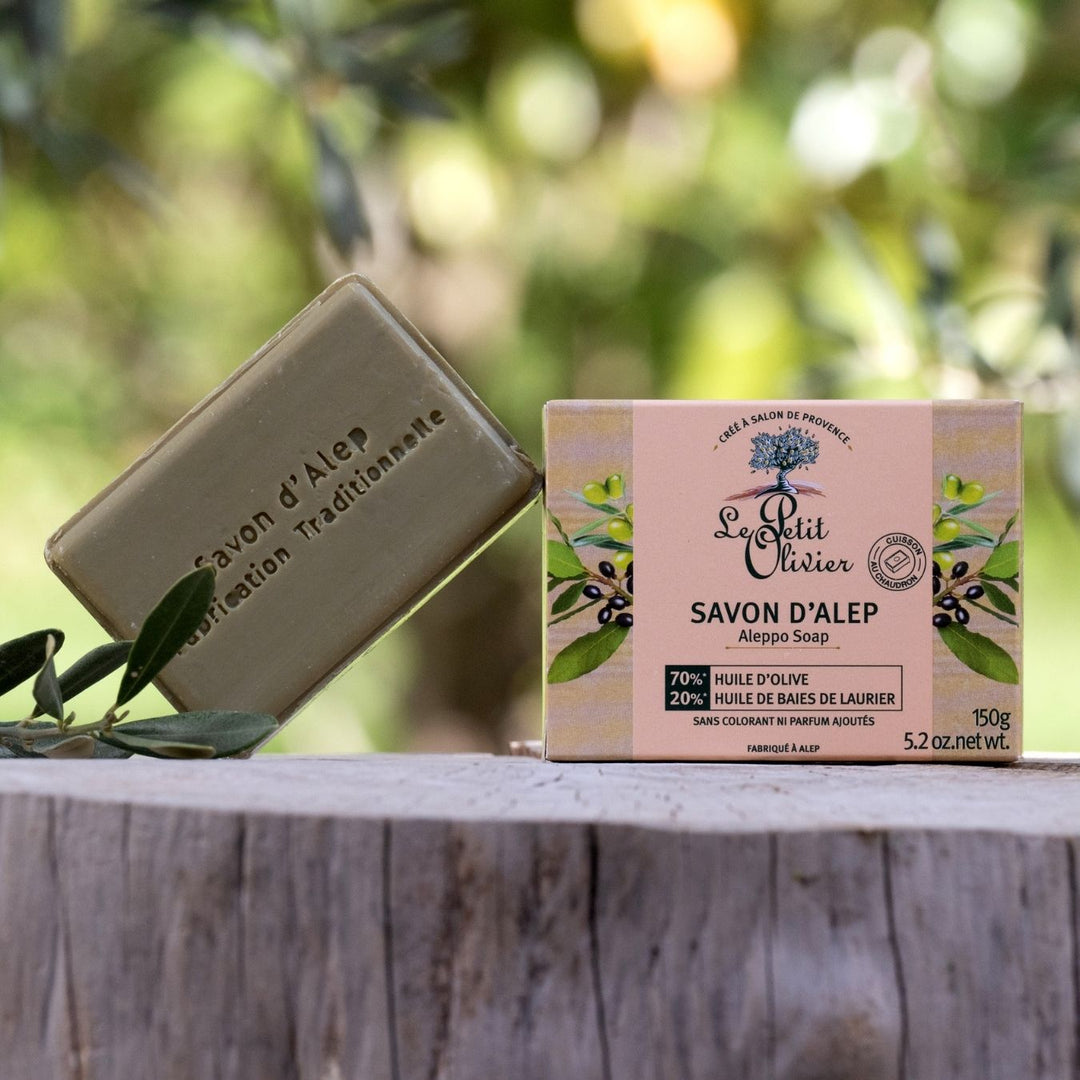 Le savon d'Alep « Le Petit Olivier », enrichi en huile d'olive et idéal pour les peaux sensibles, est mis en valeur dans son coffret fleuri, posé sur une surface en bois rustique à l'extérieur, avec en arrière-plan une végétation floue.