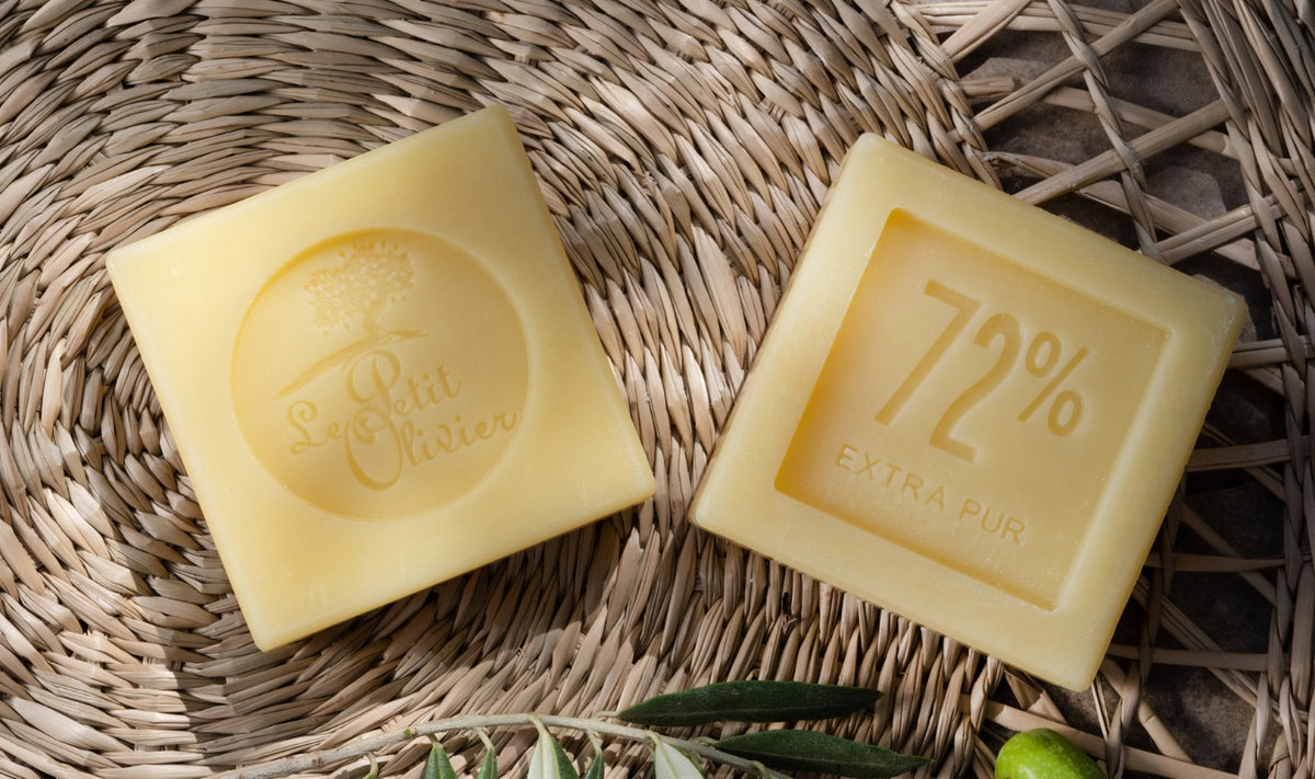 Deux pains de savon carrés reposent sur une surface tressée. L'un porte l'inscription « Le Petit Olivier » accompagnée d'un logo représentant un arbre, tandis que l'autre indique « 72 % EXTRA PUR ». Une branche d'olivier se trouve à proximité.
