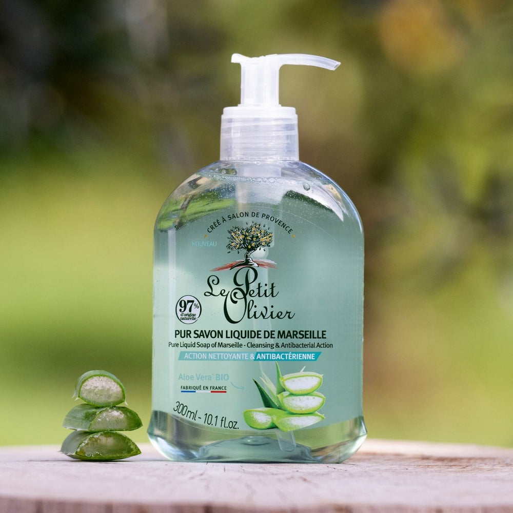 Un flacon de « Le Petit Olivier Pur Savon Liquide de Marseille - Parfum Aloe Vera » est posé à l'extérieur, à côté de trois morceaux de gel d'aloe vera empilés, avec de la verdure et un arrière-plan naturel flou qui mettent en valeur ce savon traditionnel de fabrication française.