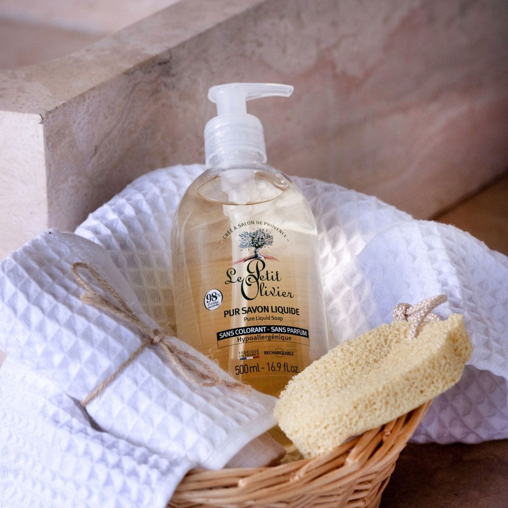 Un flacon de « Le Petit Olivier Pur Savon Liquide - Sans Parfum » repose dans un panier en osier, à côté d'une serviette blanche texturée et d'une éponge naturelle, le tout disposé sur un fond neutre.
