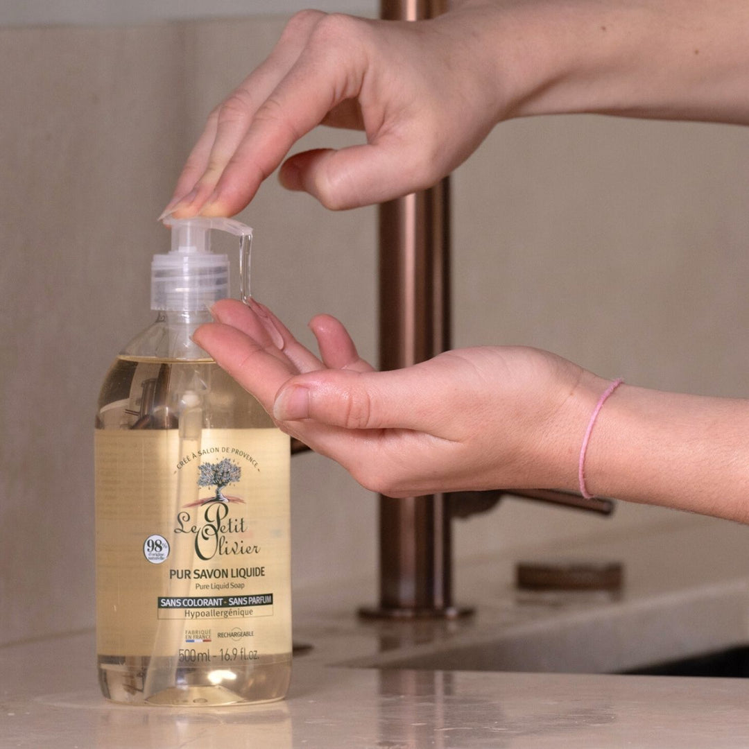 Une personne se verse du savon liquide « Le Petit Olivier Pur » – sans parfum, contenu dans un flacon transparent muni d'une pompe, dans la main, près d'un lavabo.