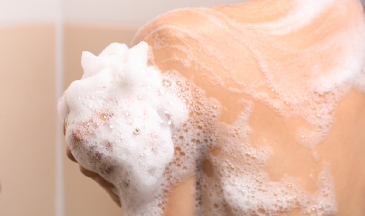 Une personne en train de se laver l'épaule avec une éponge savonneuse, recouverte d'une mousse blanche. L'image se concentre sur le haut du dos et la région de l'épaule, montrant des bulles et de la mousse de savon sur la peau.