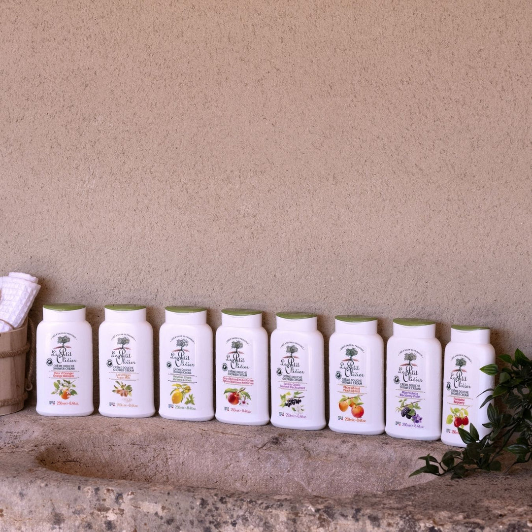 Huit flacons blancs de « Le Petit Olivier Crème Douche Jasmin Cassis », ornés chacun d'illustrations de fruits, sont alignés sur un plan de travail en pierre devant un mur beige, flanqués d'un panier contenant des serviettes pliées et d'une branche feuillue.
