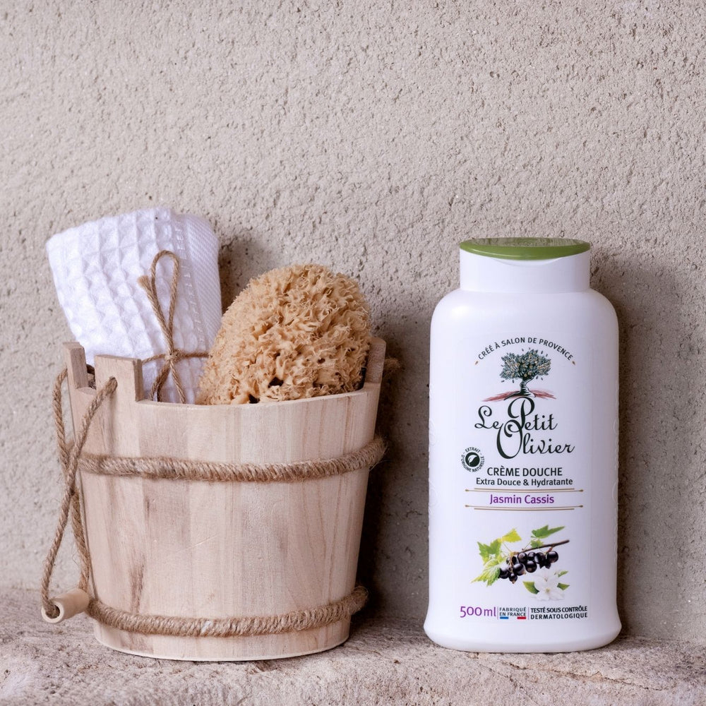 Un seau en bois contenant une serviette blanche et une éponge naturelle est posé à côté d'un flacon de « Le Petit Olivier Crème Douche Jasmin Cassis », fabriqué en France, sur fond d'un mur beige texturé.
