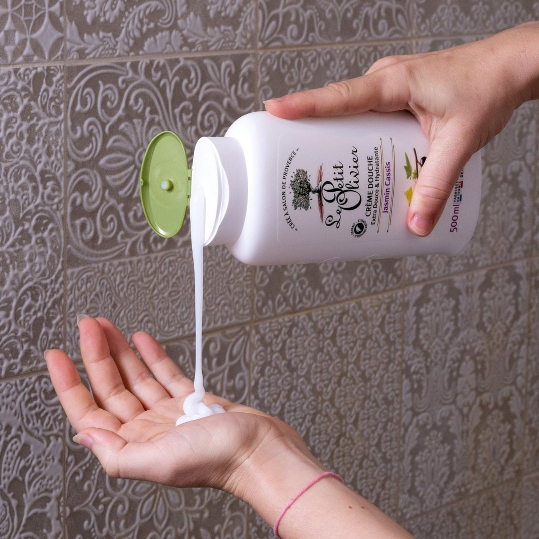 Une personne verse de la « Crème Douche Jasmin Cassis » de Le Petit Olivier, provenant d'un flacon muni d'un bouchon vert, dans sa paume ouverte, devant un mur recouvert de carrelage décoratif.