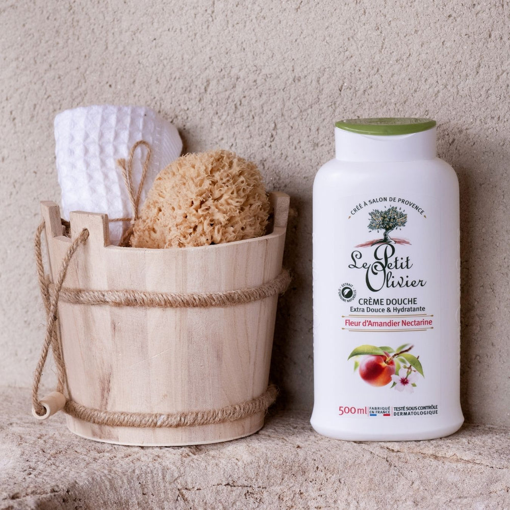 Un seau en bois contenant une serviette blanche et une éponge naturelle est posé à côté d'un flacon de « Crème Douche Fleur d'Amandier Nectarine » de Le Petit Olivier, promettant une peau hydratée sur un fond beige texturé.