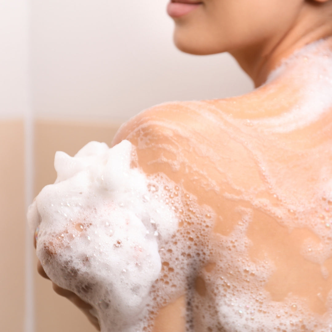 Une personne se lave l'épaule à l'aide d'une éponge de bain moussante et de la Crème Douche Fleur d'Amandier Nectarine de Le Petit Olivier, faisant naître des bulles et une mousse onctueuse sur sa peau hydratée. Son visage est partiellement visible sur un arrière-plan flou.