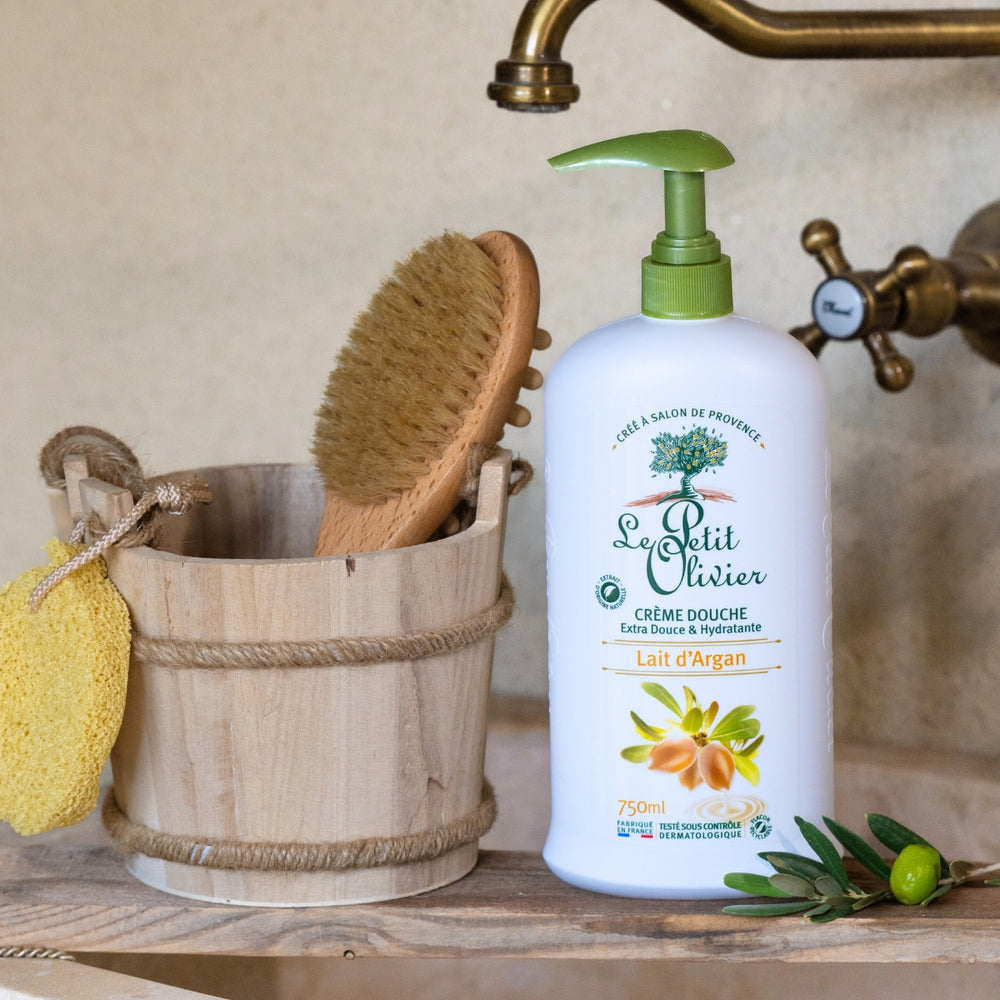 Un flacon de « Le Petit Olivier Crème Douche Lait d'Argan » est posé à côté d'un seau en bois contenant une brosse et une éponge jaune, près d'un robinet en laiton et d'un rameau d'olivier, sur le plan de toilette.