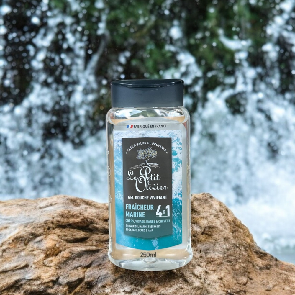 Un flacon de gel douche pour homme « Le Petit Olivier – Vivifiant – Fraîcheur Marine » repose sur un rocher, avec une cascade et de la végétation en arrière-plan, le tout flou. Son étiquette bleue et noire comporte un texte en blanc et bleu clair.