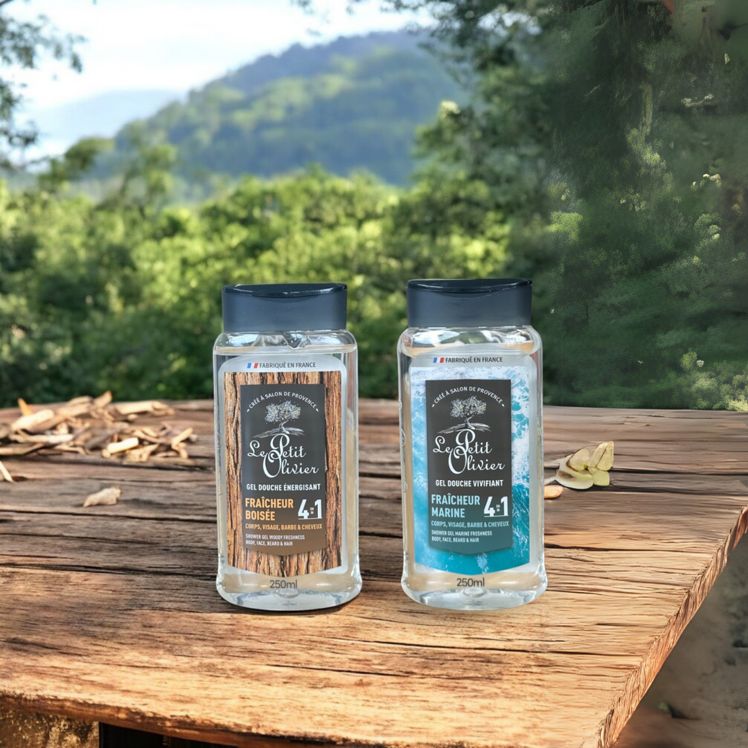 Deux flacons de gel douche pour homme « Le Petit Olivier » Énergisant, l'un au parfum « Fraîcheur Boisée » et l'autre au parfum « Fraîcheur Marine », sont posés sur une surface en bois à l'extérieur, avec en arrière-plan des arbres verts et des montagnes floues.