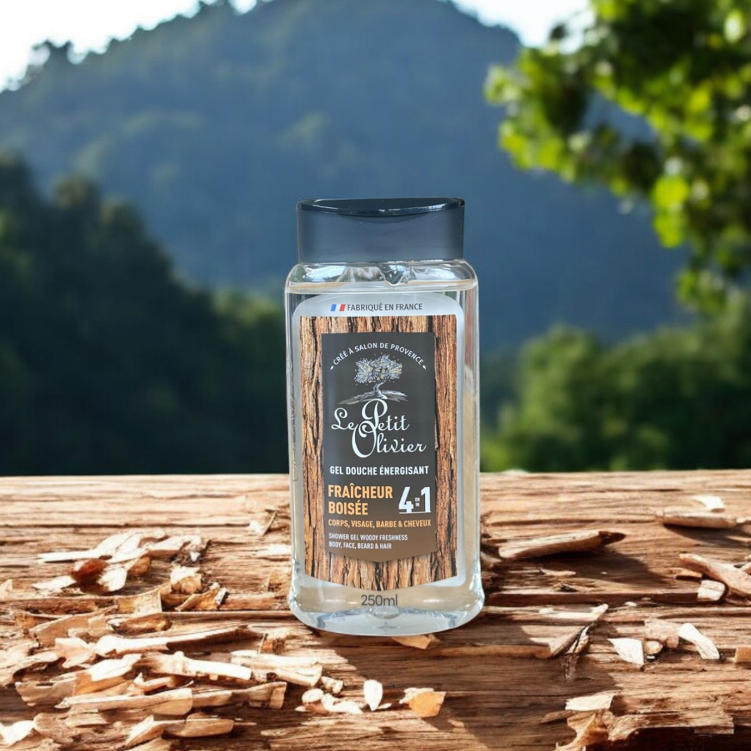 Un flacon de gel douche pour homme « Le Petit Olivier » Énergisant - Fraîcheur Boisée repose sur une surface en bois parsemée de copeaux, avec en arrière-plan une montagne boisée aux contours flous.