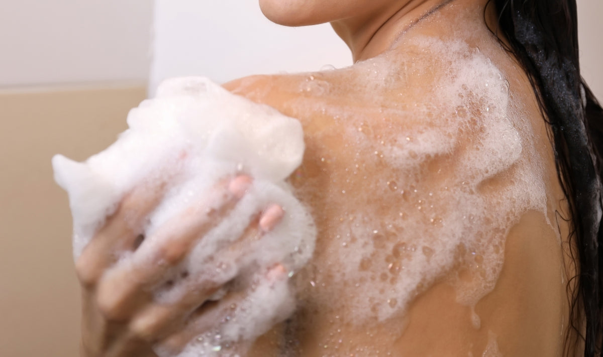 Une personne aux cheveux mouillés se lave l'épaule à l'aide d'une éponge savonneuse ; des bulles et de la mousse recouvrent sa peau. La photo met en avant l'épaule, la main et le haut du dos.