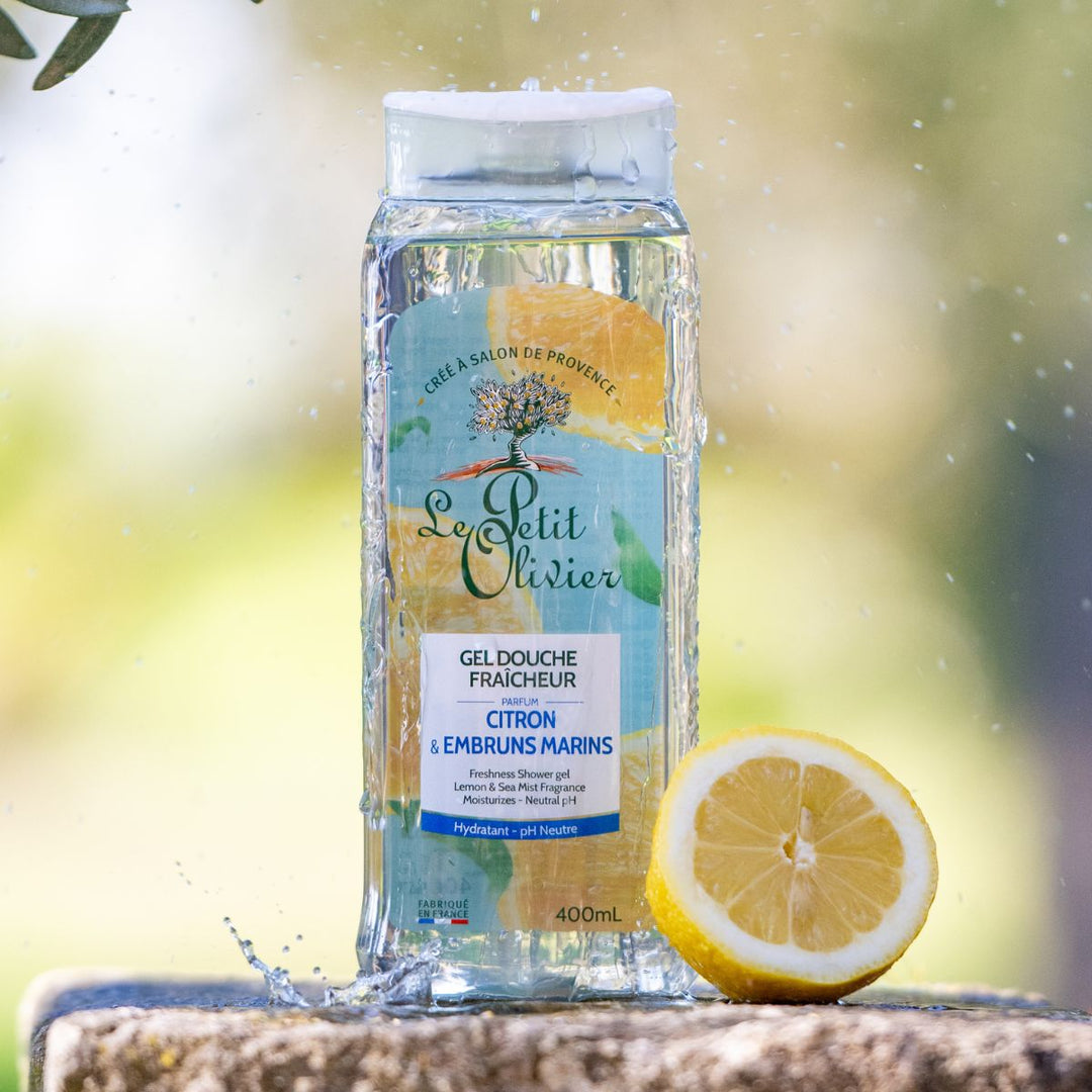 Une bouteille de gel douche « Fraîcheur » de Le Petit Olivier est posée sur un rebord en pierre à l'extérieur, avec des éclaboussures d'eau à proximité et un citron coupé en deux à côté. Fabriqué en France, la végétation apparaît légèrement floue en arrière-plan.