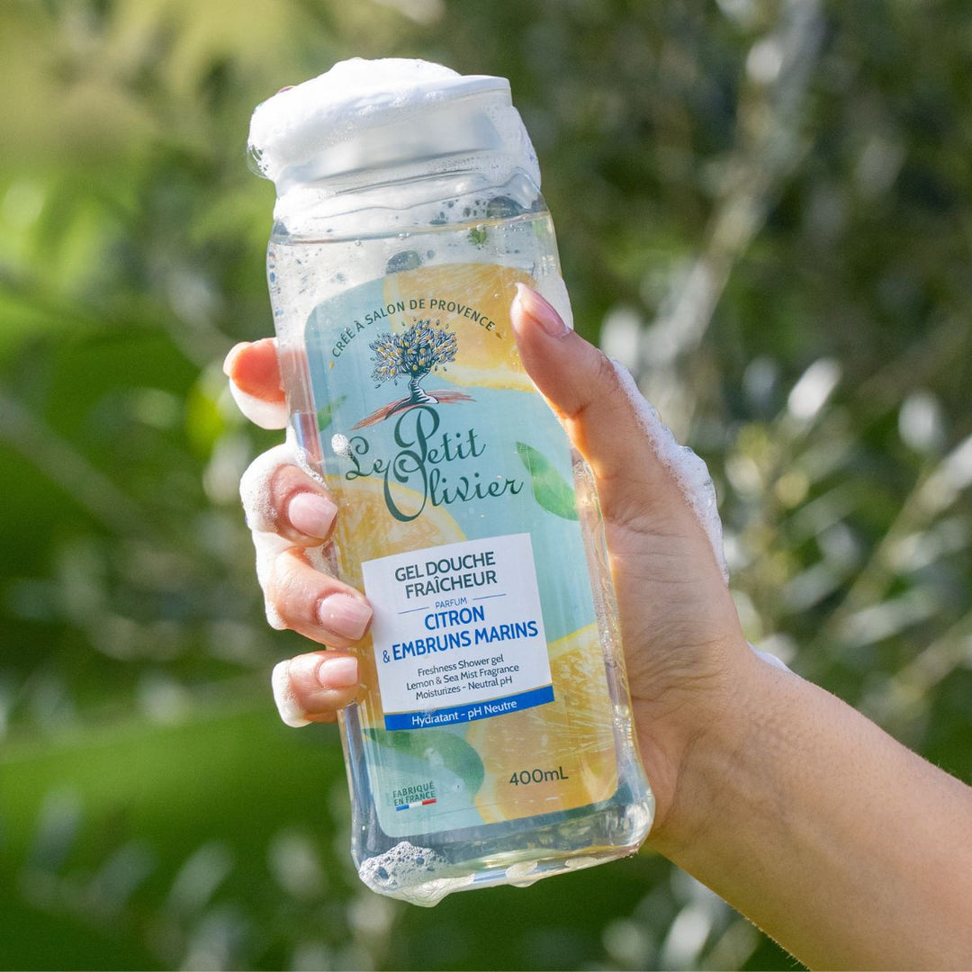 Une main savonneuse tient un flacon transparent de gel douche « Fraîcheur » de Le Petit Olivier. L'étiquette porte la mention « fabriqué en France ». L'arrière-plan, d'un vert feuillu et légèrement flou, évoque la fraîcheur.