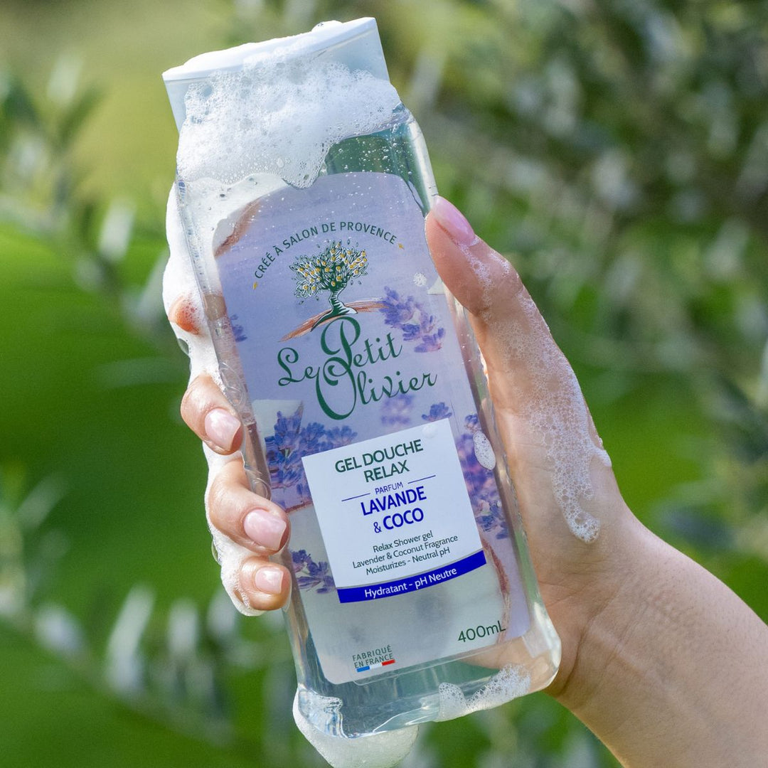 Une main savonneuse tient un flacon de gel douche « Le Petit Olivier Relax » devant un décor extérieur vert et flou.