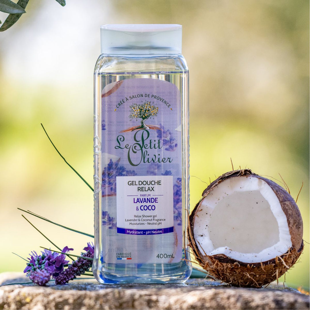 Un flacon de gel douche « Le Petit Olivier Relax » est posé à l'extérieur, à côté d'une noix de coco coupée en deux et de brins de lavande, sur un fond vert flou.