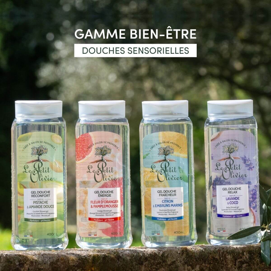 Quatre flacons de gel douche « Le Petit Olivier Relax » sont présentés en extérieur sur un fond naturel. Chaque flacon, au pH neutre, arbore une étiquette aux couleurs vives et un parfum distinct : pistache-amande, néroli-pamplemousse, citron-brise marine et lavande-noix de coco.