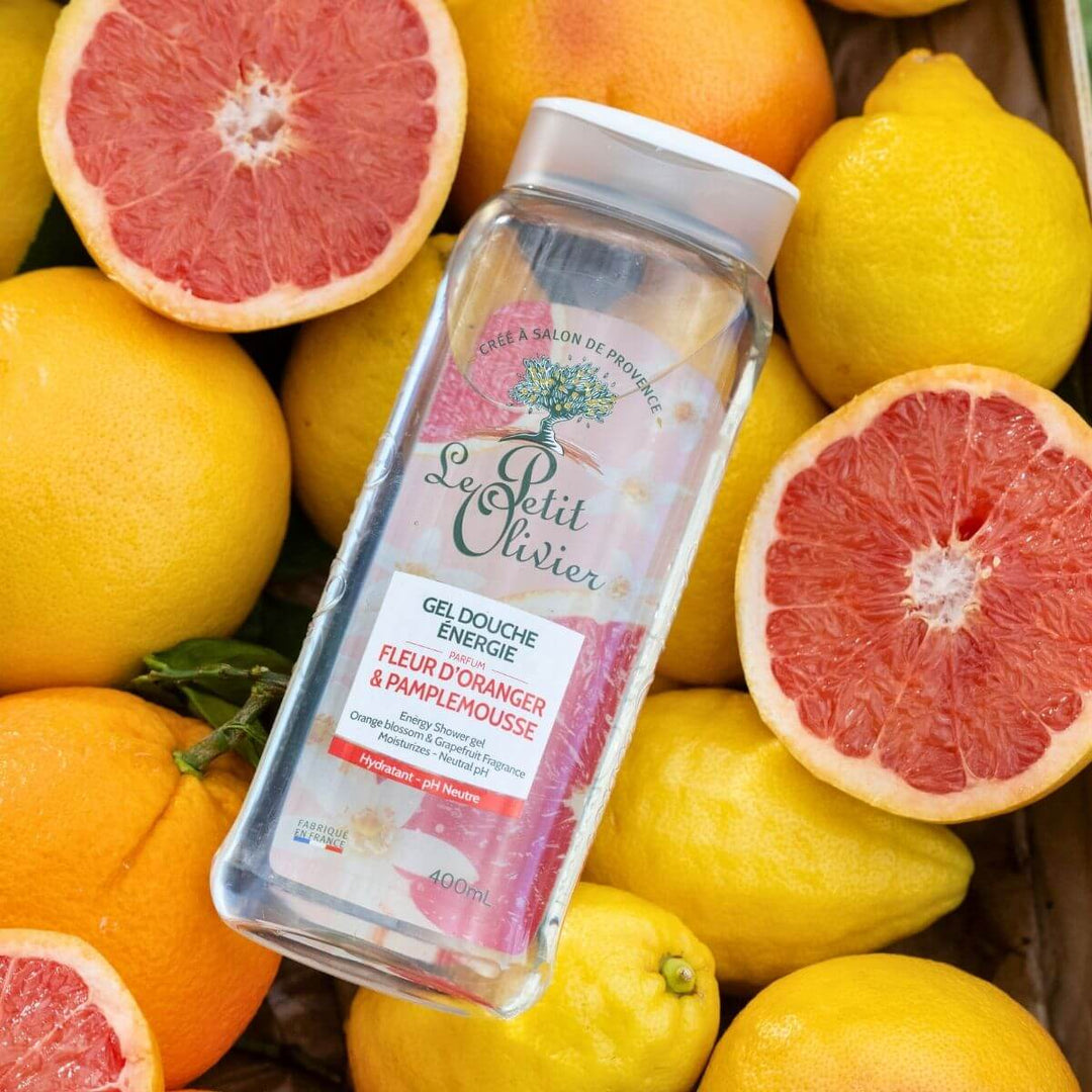 Un flacon de gel douche Énergie « Le Petit Olivier » trône au milieu d'oranges, de pamplemousses et de citrons frais. L'étiquette arbore des illustrations aux couleurs vives représentant des agrumes, mettant en valeur le mélange énergisant d'orange et de pamplemousse.