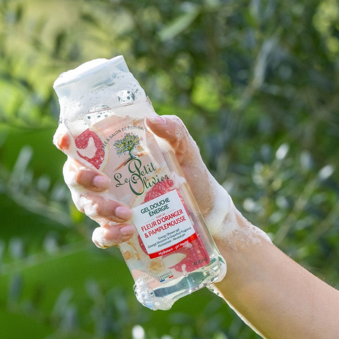 Une main savonneuse tient un flacon de gel douche « Le Petit Olivier Énergie », sur fond flou représentant un paysage extérieur verdoyant.