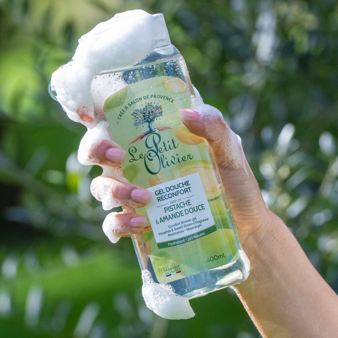 Une main recouverte de mousse de savon tient un flacon de Gel Douche Réconfort de Le Petit Olivier, sur fond flou représentant un paysage extérieur verdoyant.