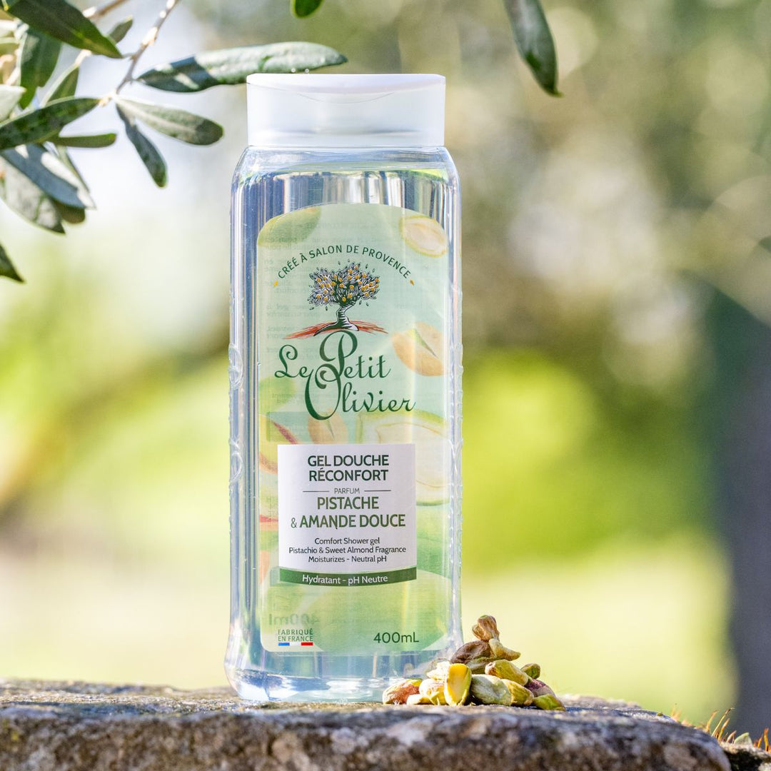 Une bouteille de gel douche « Le Petit Olivier Réconfort » trône sur une surface en pierre à l'extérieur, au milieu de pistaches, sur un fond vert baigné de soleil — l' cadre idéal pour une expérience apaisante avec ce gel douche.