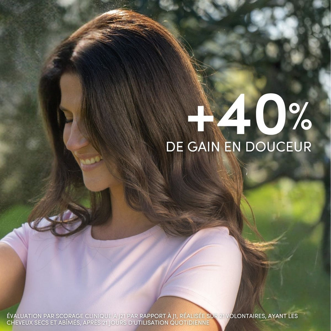 Une femme souriante aux longs cheveux bruns et brillants se tient à l'extérieur, baignée par la lumière du soleil, vêtue d'un t-shirt rose pâle. Crème de Soin Cheveux Secs et Abîmés de Le Petit Olivier aux huiles d'olive, de karité et d'argan : +40 % de douceur.