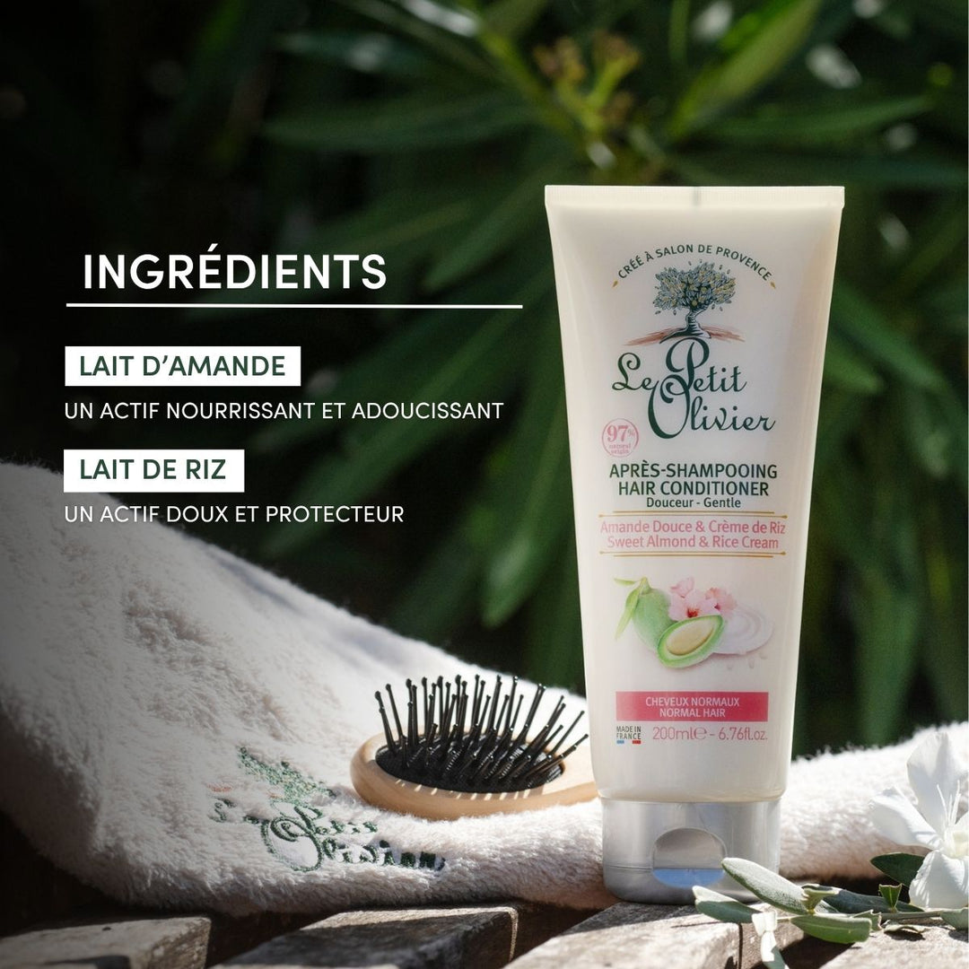 Un tube d'après-shampoing « Le Petit Olivier » pour cheveux normaux repose sur une serviette, à côté d'une brosse à cheveux noire, baigné par la lumière du soleil. La marque française met en avant ses ingrédients nourrissants : le lait d'amande douce et le lait de riz.