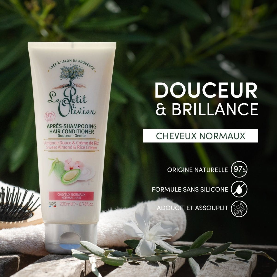 Un tube d’après-shampoing « Le Petit Olivier » pour cheveux normaux est posé à la verticale sur une serviette blanche à l’extérieur, entouré de feuillage vert. L’emballage met en avant le lait d’amande douce et est destiné aux cheveux normaux.