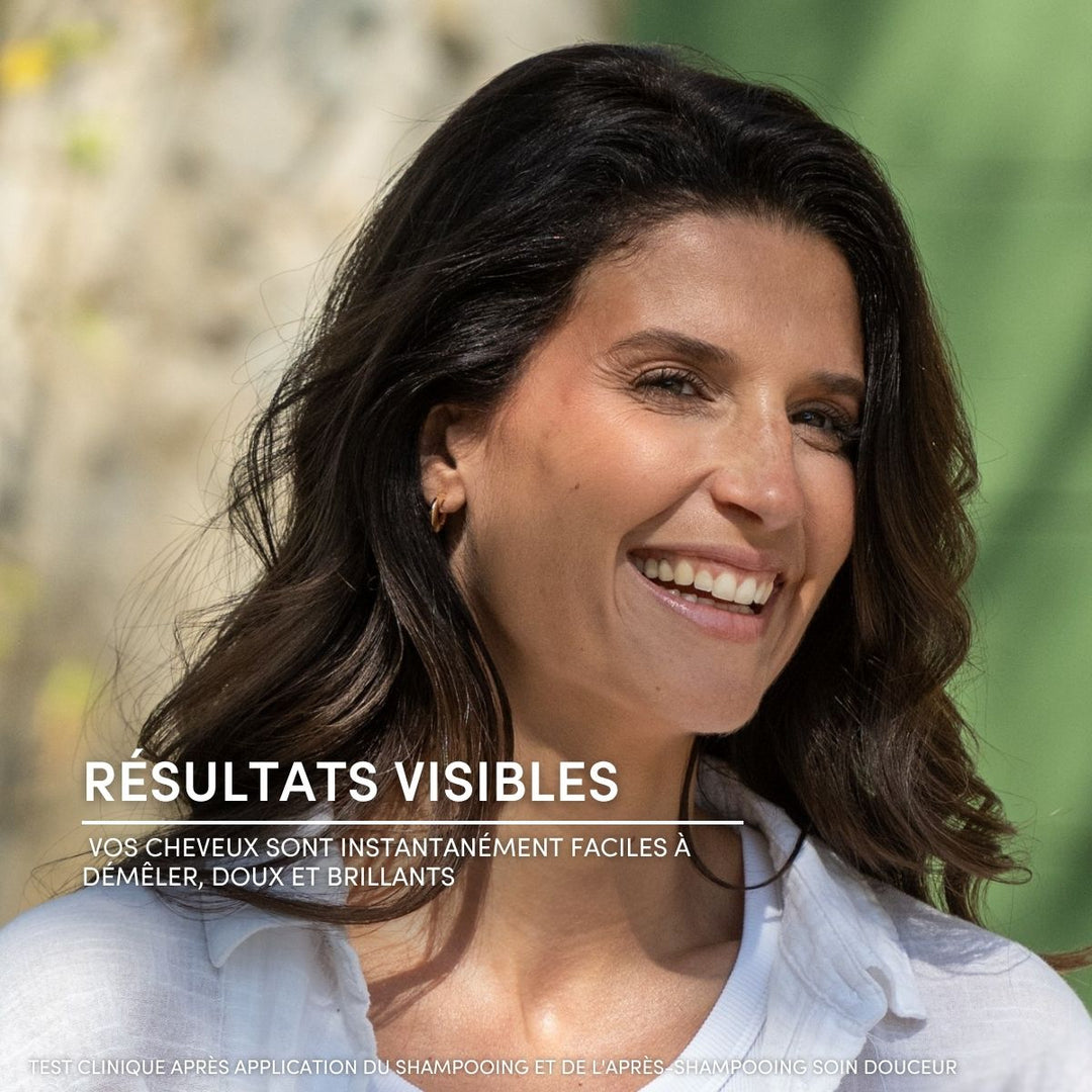 Une femme souriante aux cheveux bruns ondulés se tient à l'extérieur, baignée par la lumière du soleil, vêtue d'une chemise blanche. Un texte en français souligne que l'après-shampoing Le Petit Olivier pour cheveux normaux rend les cheveux lisses, brillants et faciles à démêler après utilisation.