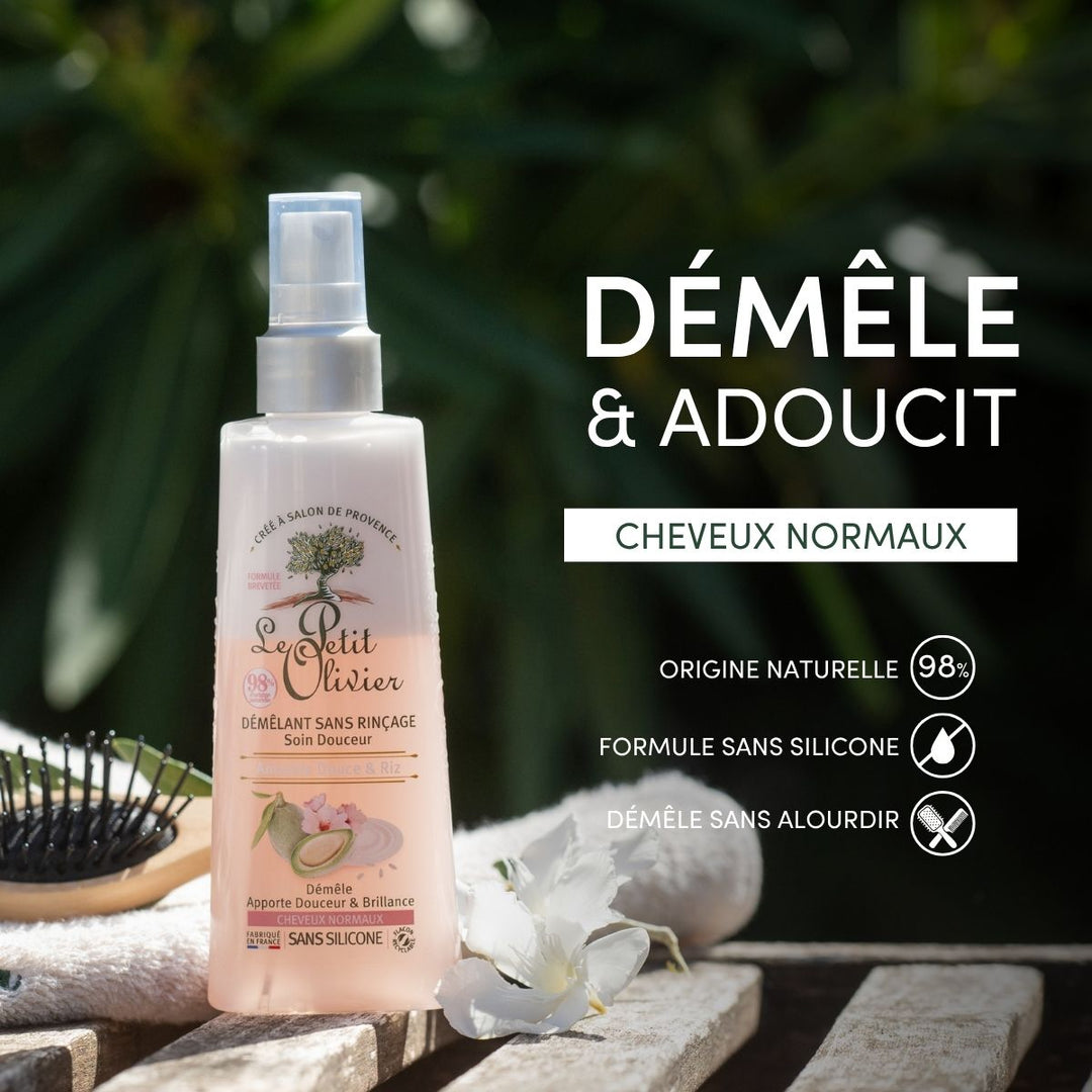 Un flacon de « Le Petit Olivier Spray Démêlant Sans Rinçage Cheveux Normaux » est posé à l'extérieur, à côté d'une brosse à cheveux et de fleurs. L'étiquette précise son origine naturelle et sa formule sans silicone. Le texte indique : « Démêle & Adoucit Cheveux Normaux ».