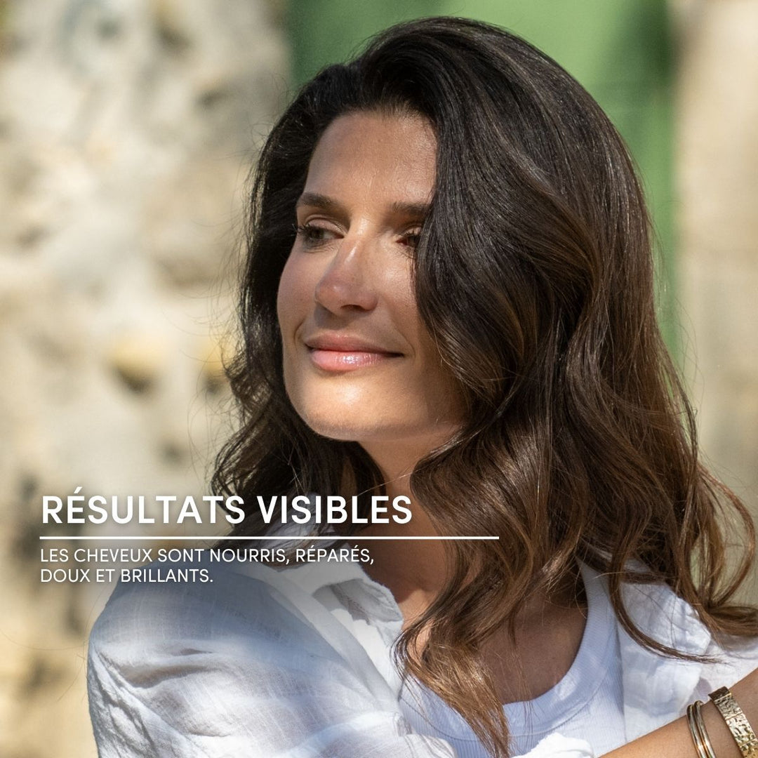 Une femme aux longs cheveux bruns ondulés et brillants sourit en extérieur, vêtue d'un haut blanc. Texte en français : RÉSULTATS VISIBLES. Le Petit Olivier Spray démêlant sans rinçage pour cheveux normaux, sans silicone et idéal pour les cheveux normaux.