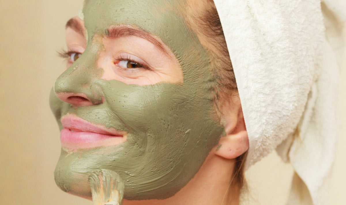 Une personne, la tête enveloppée d'une serviette, applique un masque facial vert à l'aide d'un pinceau, en souriant doucement.