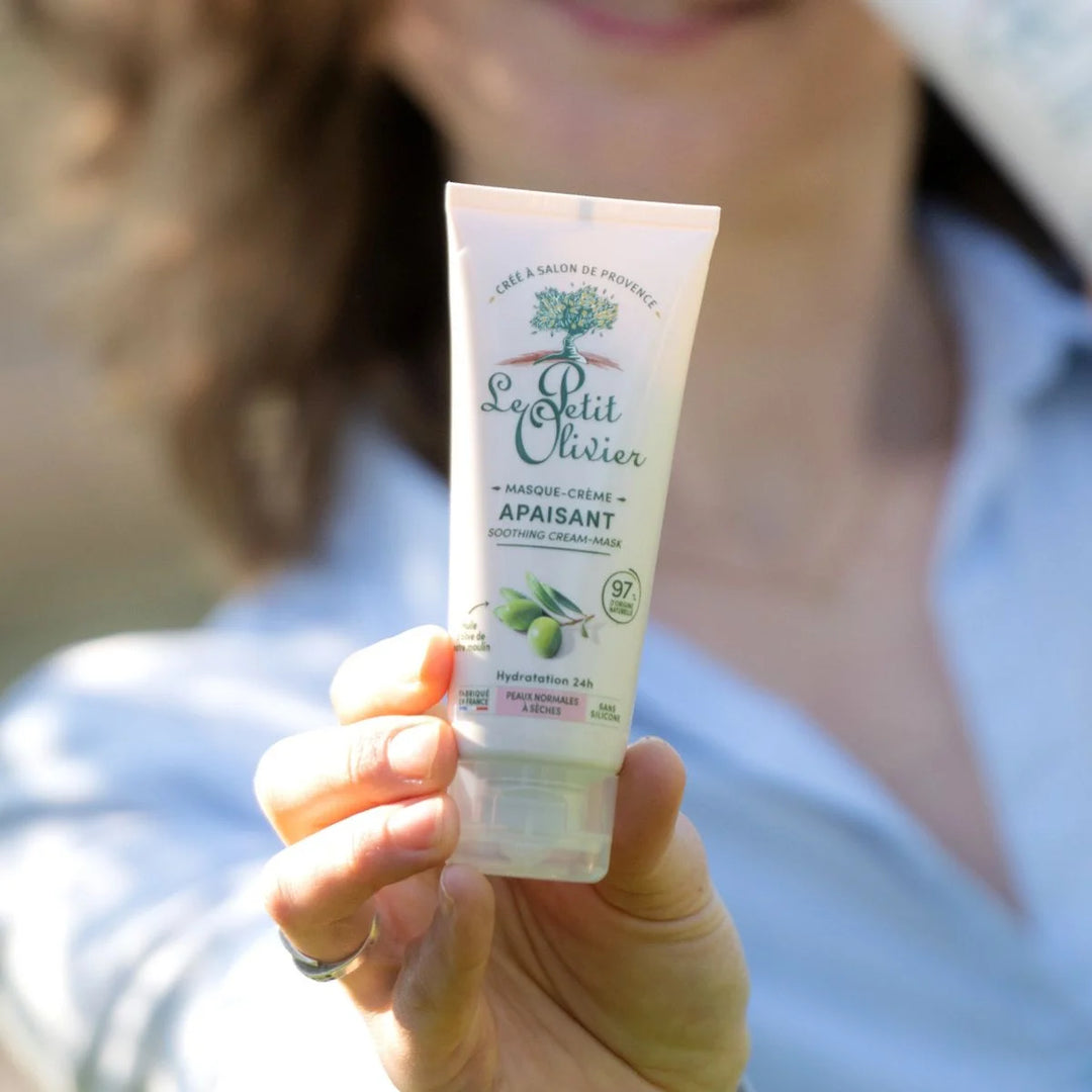 Une personne tient un tube de « Le Petit Olivier - Masque-crème apaisant » dont l'étiquette représente des olives. L'arrière-plan est flou, mettant ainsi le produit en valeur.
