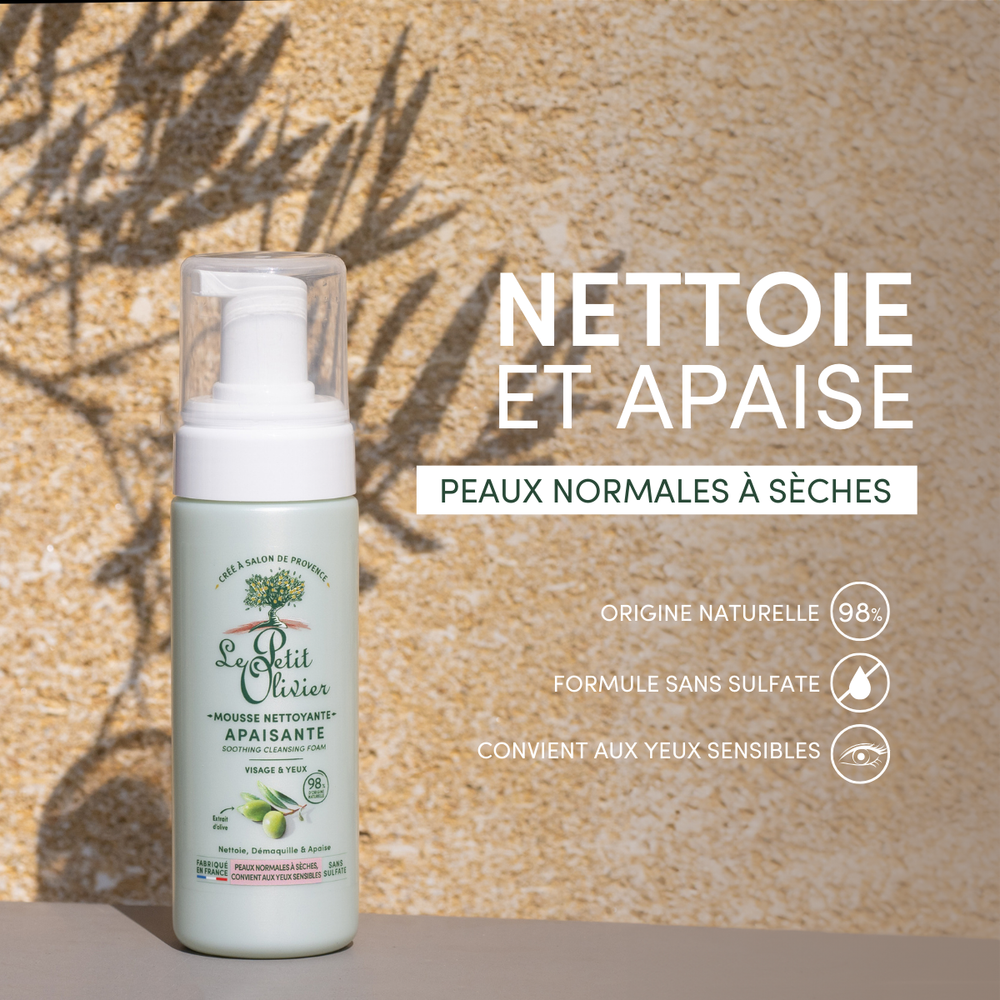 A bottle of Mousse Nettoyante Visage Apaisante stands on a beige surface with shadowed leaves behind. French text highlights its olive extract, sulfate-free formula, and gentle cleansing for dry skin and sensitive eyes.