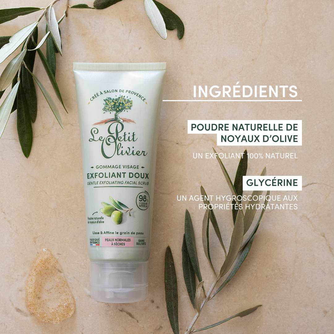 Un tube de « Le Petit Olivier Gommage Visage Exfoliant Doux » repose sur une surface beige parsemée de feuilles d'olivier. Le texte met en avant la poudre naturelle de noyau d'olive et la glycérine comme ingrédients clés.