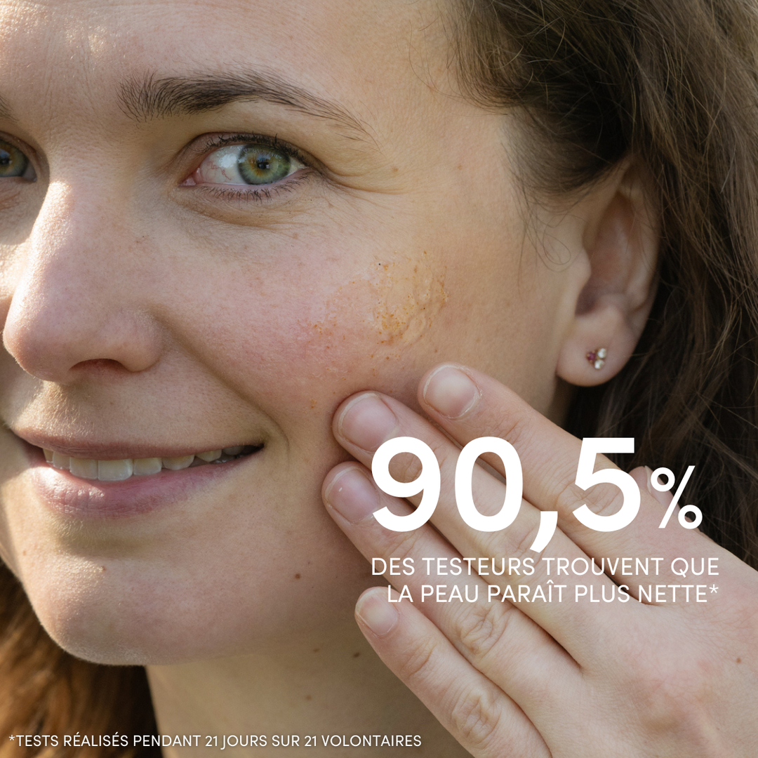 Une femme à la peau claire et aux yeux bleus sourit tout en appliquant le « Gommage Visage Exfoliant Doux » de Le Petit Olivier sur sa joue droite. Le texte en français indique : « 90,5 % des testeurs trouvent que la peau paraît plus nette. ».