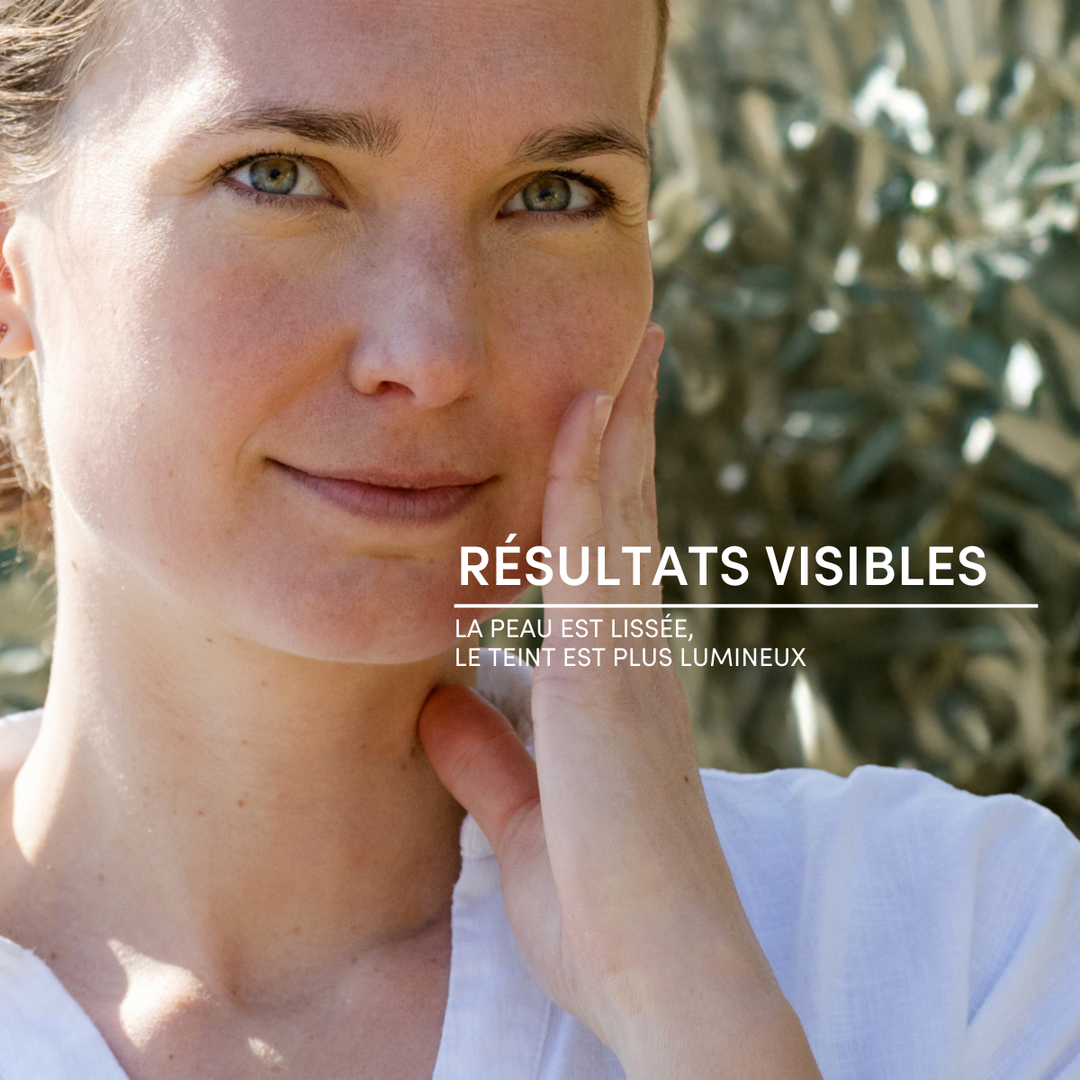 Une femme vêtue d'une chemise blanche, arborant un doux sourire, se touche la joue. Texte en français : RÉSULTATS VISIBLES. LA PEAU EST LISSÉE, LE TEINT PLUS LUMINEUX grâce au Gommage Visage Exfoliant Doux de Le Petit Olivier. Un feuillage vert flou apparaît en arrière-plan.
