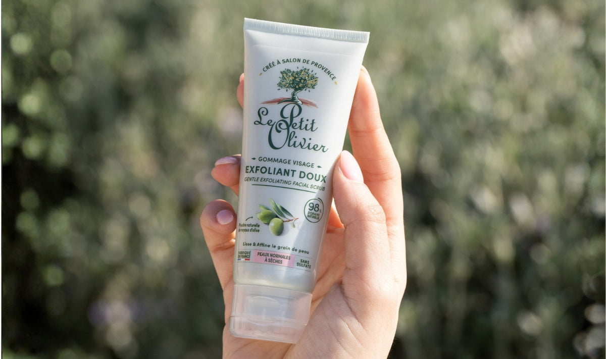 Une main, dont les ongles sont vernis d'un violet clair, tient un tube de gommage visage « Exfoliant Doux » de Le Petit Olivier, sur un fond vert flou représentant un extérieur.