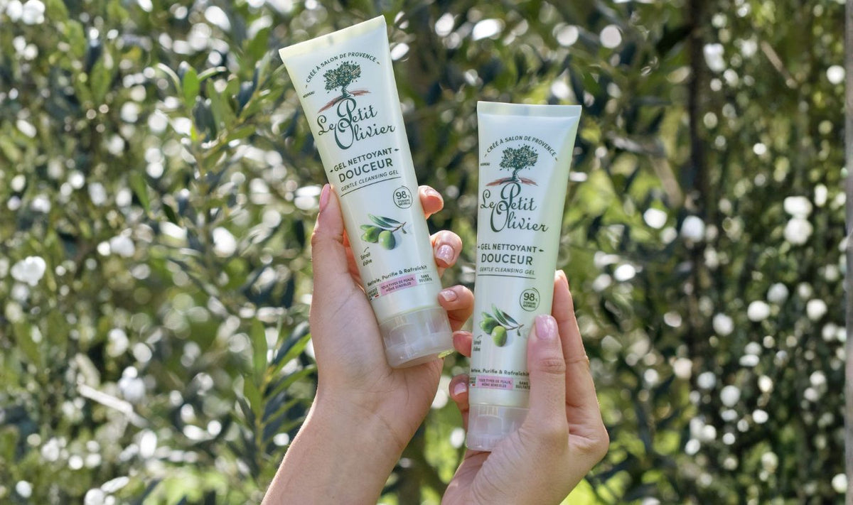Deux mains tiennent des tubes de gel nettoyant doux « Le Petit Olivier » à l'extrait d'olive, sur fond de branches verdoyantes et de rayons de soleil.