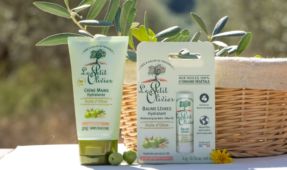 Deux produits de soin à l'huile d'olive de la marque Le Petit Olivier sont présentés devant un panier en osier et des branches d'olivier : un tube de crème pour les mains et un baume à lèvres emballé, tous deux ornés de touches de vert et d'illustrations d'olives.