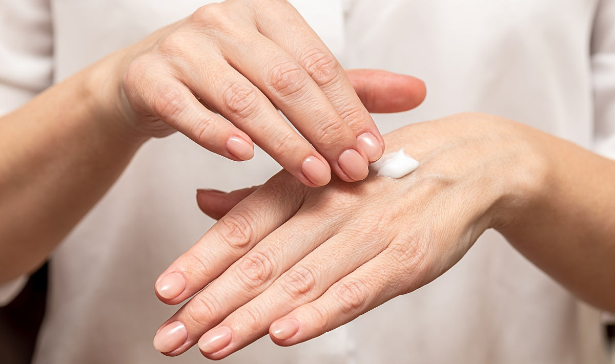 Une personne applique une lotion blanche sur le dos de sa main et la fait pénétrer en la frottant avec ses doigts. L'arrière-plan est flou ; seules les mains et une partie d'une chemise blanche sont visibles.