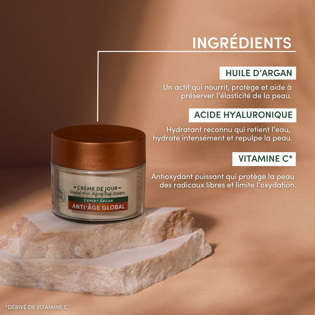 Un pot de « Crème de Jour Anti-Âge Hydratante » de Le Petit Olivier repose sur une surface beige, avec les ingrédients mis en avant en français : huile d'argan, acide hyaluronique et vitamine C*. Fond brun crémeux, ombres douces et pierres à côté du pot.