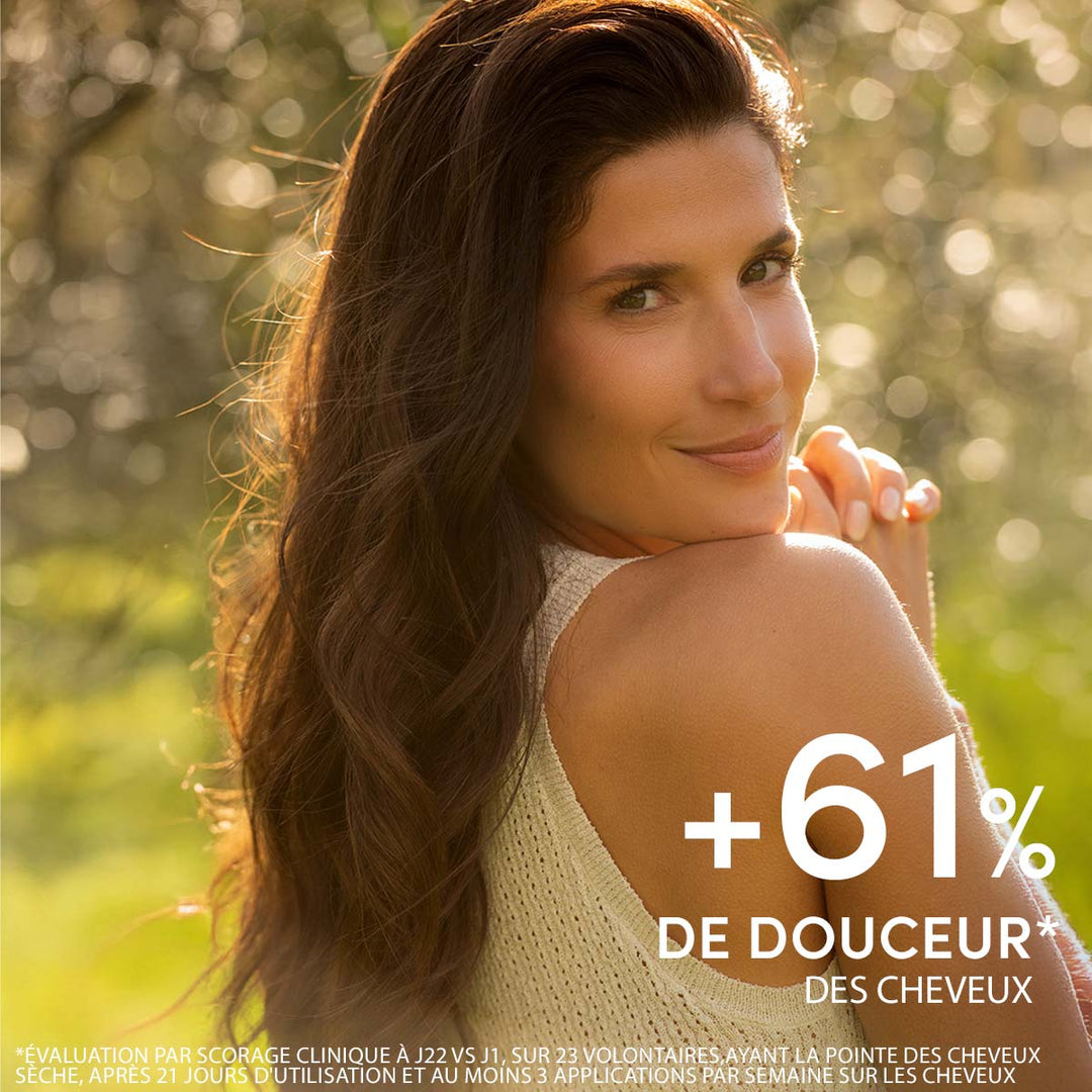 Une femme aux longs cheveux bruns ondulés sourit en plein air. Le texte indique « +61 % de douceur des cheveux », soulignant les bienfaits des huiles précieuses. Produit : Huile sèche sublimatrice pour les cheveux, le corps et le visage de Le Petit Olivier. Une note en français mentionne une évaluation capillaire.