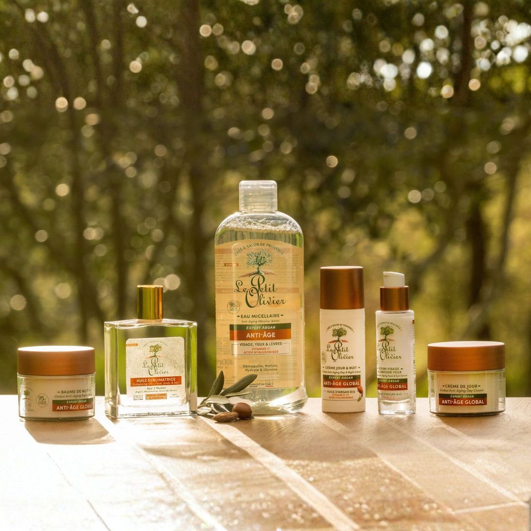 L'« Huile Sèche Sublimatrice » de Le Petit Olivier, destinée aux cheveux, au corps et au visage, est présentée sur un support en bois baigné par la lumière naturelle, au milieu des arbres, ce qui met en valeur ses huiles précieuses et ses bienfaits hydratants pour la peau.