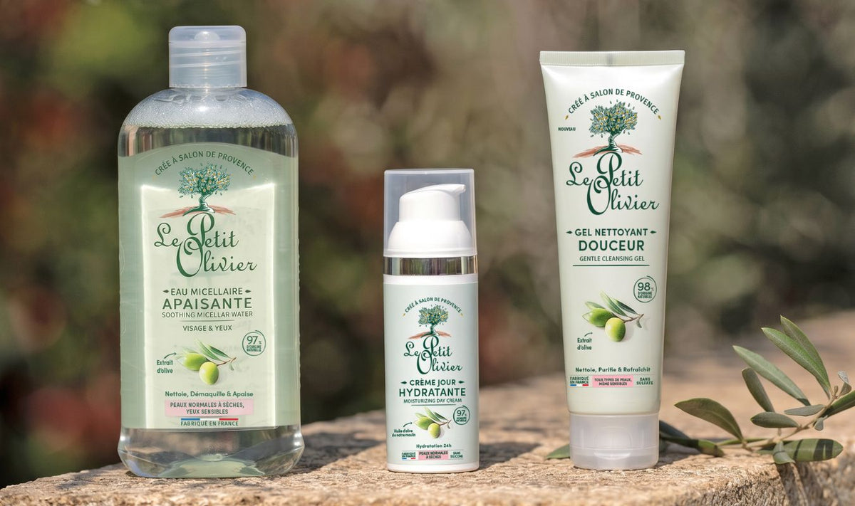 Trois produits de soin « Le Petit Olivier » — une eau micellaire, une crème hydratante et un gel nettoyant doux — sont présentés en extérieur sur une surface en pierre, avec un arrière-plan naturel flou.