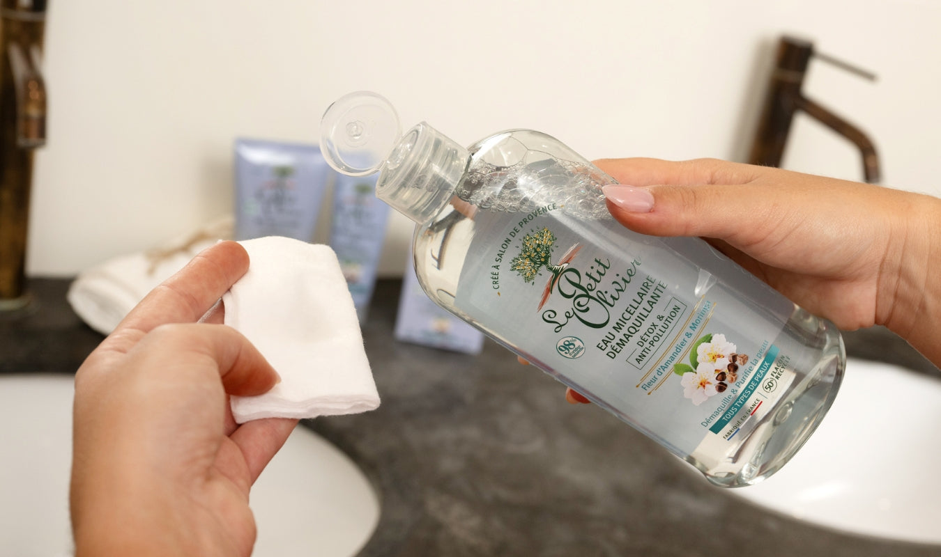 Une personne verse de l'eau micellaire démaquillante d'un flacon sur un coton, sur le plan de travail d'une salle de bains où l'on aperçoit, en arrière-plan, un lavabo et des produits de beauté.